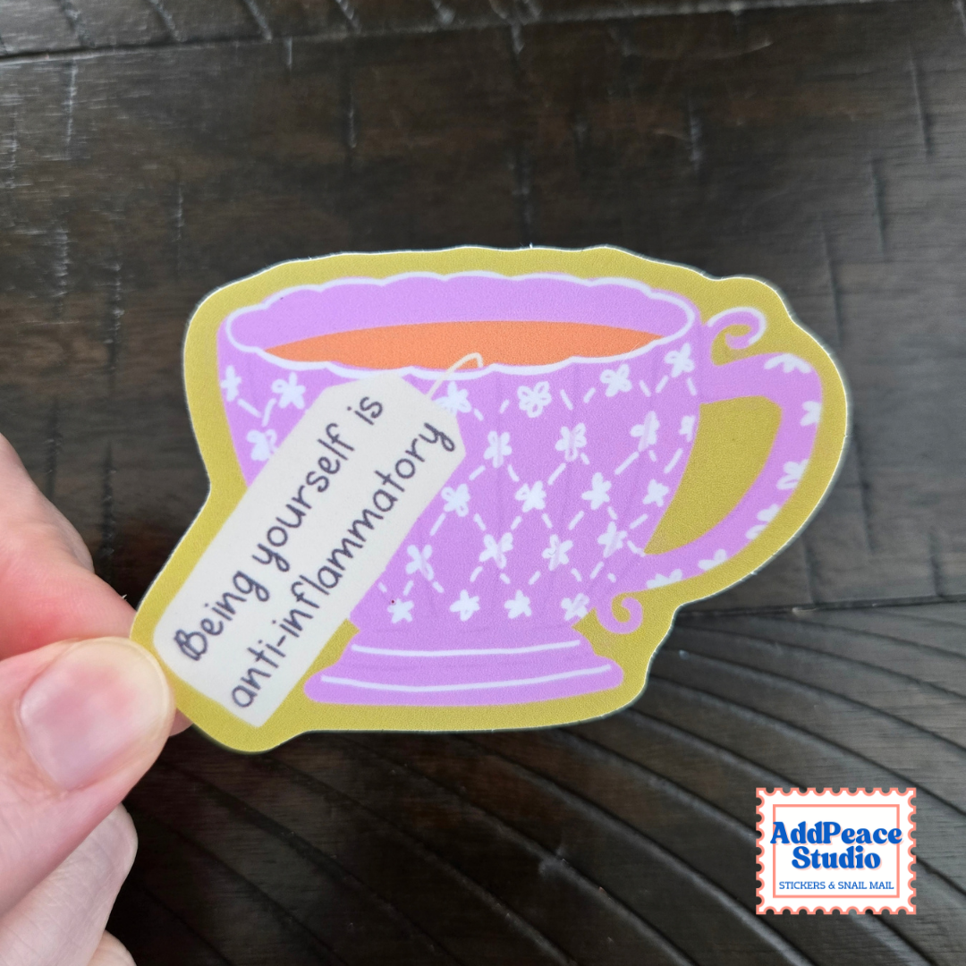 A hand holds a die-cut sticker shaped like a purple floral teacup with a yellow border and a small hanging tag that reads, 'Being yourself is anti-inflammatory.' The sticker is photographed over a dark wooden surface, with an AddPeace Studio logo in the bottom-right corner.