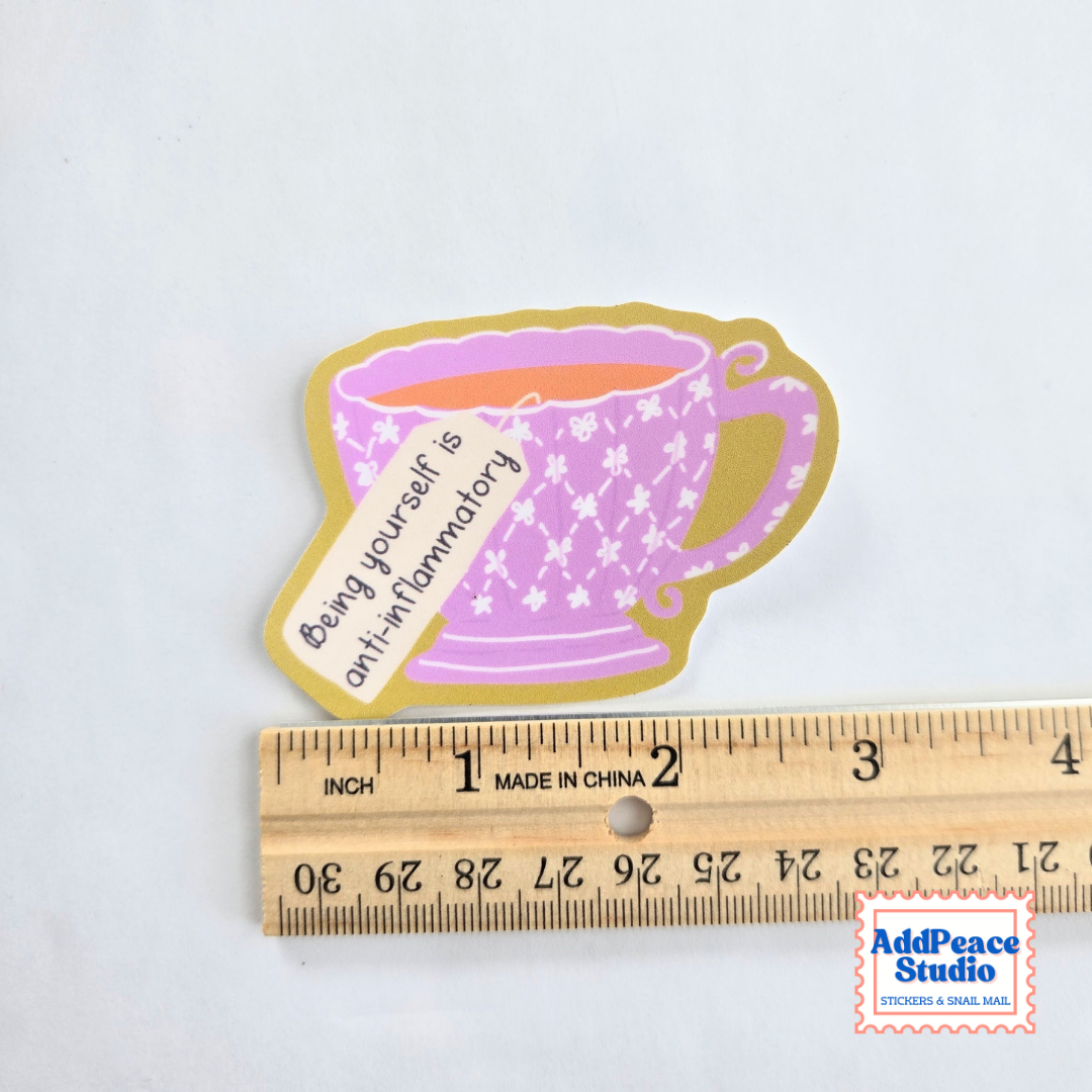 A die-cut sticker shaped like a pink patterned teacup with a gold border, placed above a wooden ruler to show scale. The sticker includes a teabag tag with the text 'Being yourself is optional.