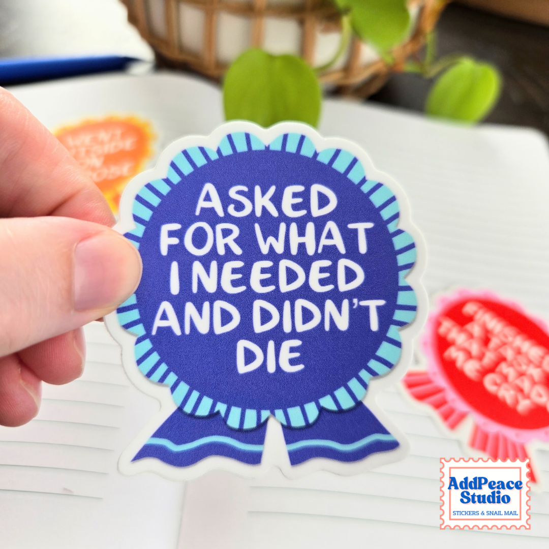 A hand holds a die-cut sticker shaped like a blue ribbon badge with the text 'ASKED FOR WHAT I NEEDED AND DIDN'T DIE' in white. The sticker is shown against a blurred desk background with other colorful stickers and a plant.