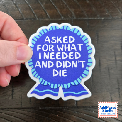 A hand holds a die-cut sticker shaped like a blue ribbon award. The sticker has white text reading 'ASKED FOR WHAT I NEEDED AND DIDN'T DIE' on a blue background with light blue stripes.