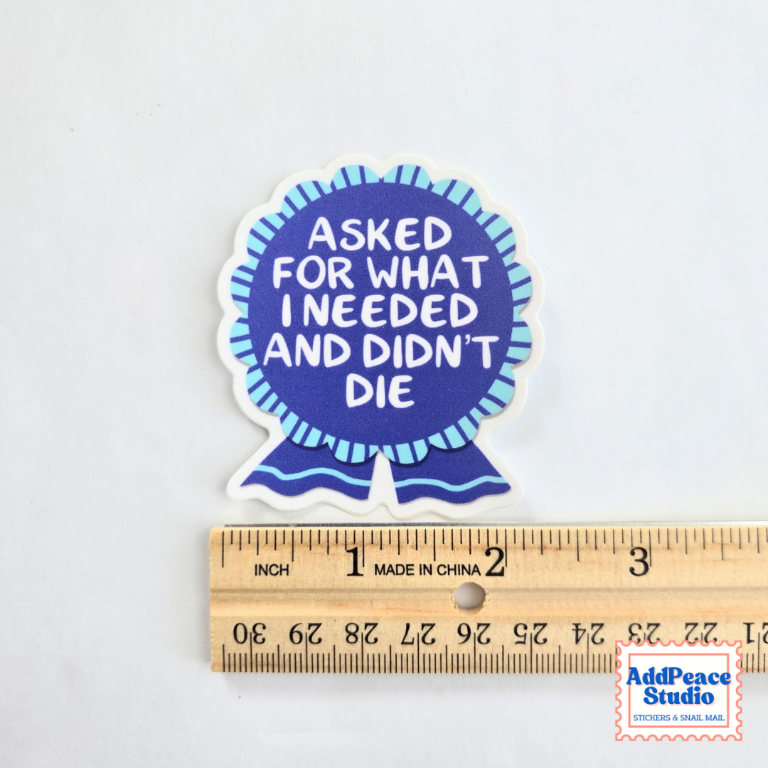 A die-cut sticker shaped like a blue ribbon badge with the text 'ASKED FOR WHAT I NEEDED AND DIDN'T DIE' in white, shown above a wooden ruler for scale on a white background.