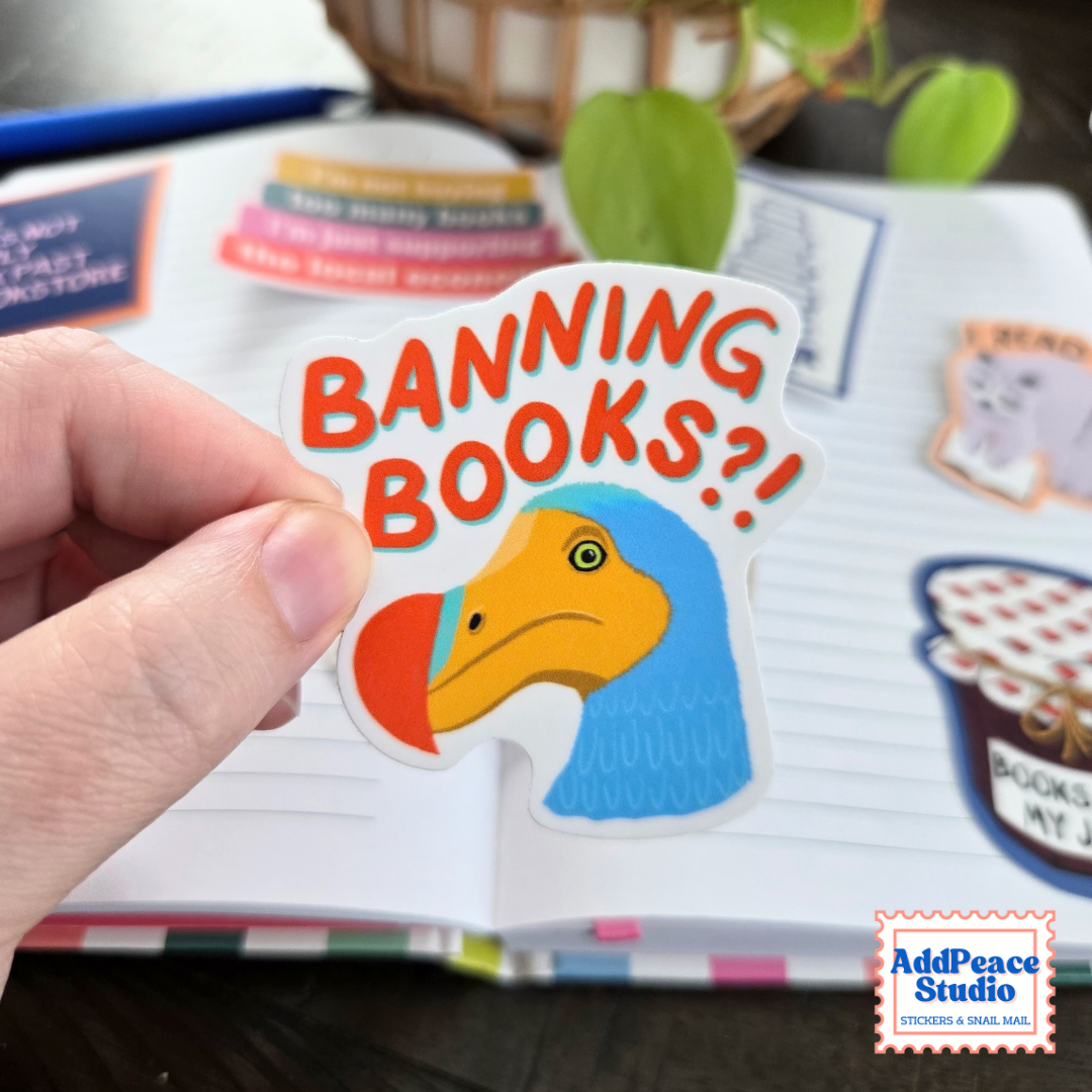 A hand holds a die-cut sticker featuring a blue and yellow Dodo bird with the text 'BANNING BOOKS?!' in orange-red letters, set against an open notebook with other stickers.