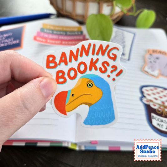 A hand holds a die-cut sticker featuring a blue and yellow Dodo bird with the text 'BANNING BOOKS?!' in orange-red letters, set against an open notebook with other stickers.