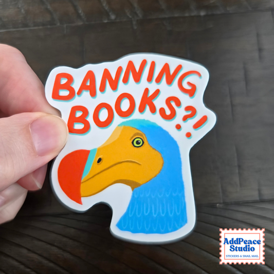 A hand holds a die-cut sticker featuring a dodo bird illustration and the text 'BANNING BOOKS?!' in orange letters with a blue shadow effect, against a dark wood background. An 'AddPeace Studio' logo is visible in the bottom right corner.