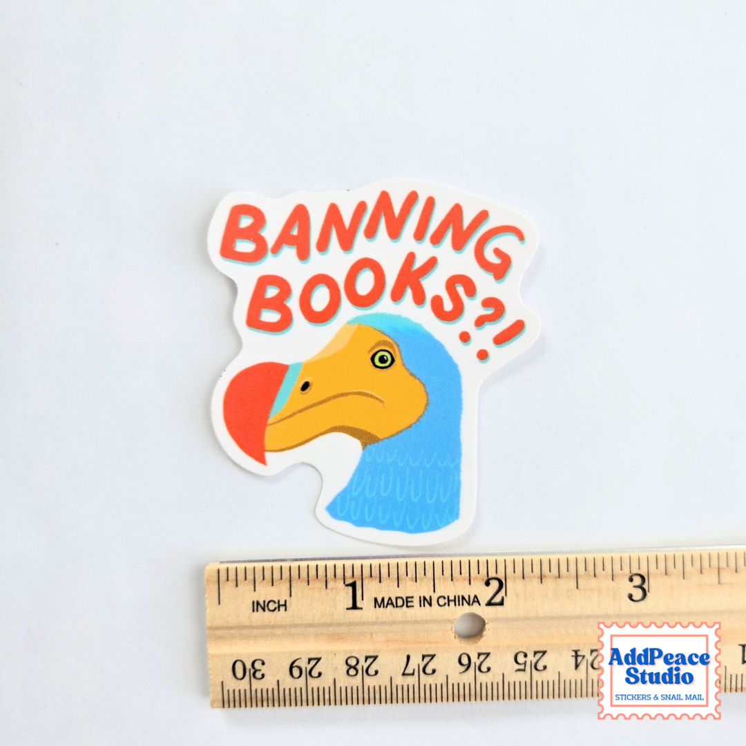 A die-cut sticker featuring a cartoon Dodo bird with a blue body, yellow beak, and red beak, with the text 'BANNING BOOKS?!' in red-orange lettering. The sticker is displayed above a wooden ruler to show its size, on a plain white background.
