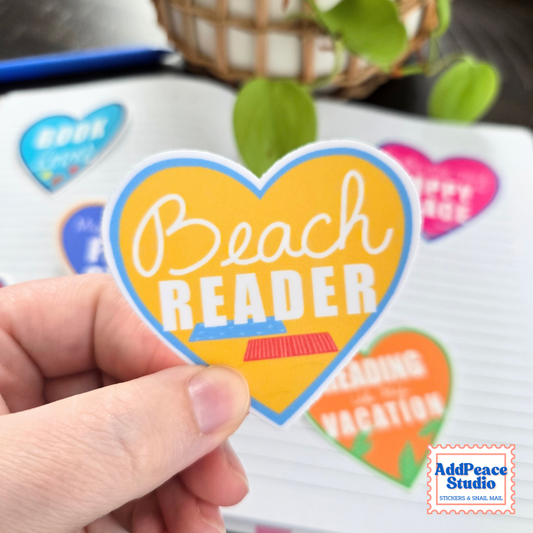 A hand holds a heart-shaped sticker that reads 'Beach Reader' in white script and block letters on a yellow background with a blue border. The scene is a styled flat-lay with other colorful stickers on a notebook and blurred green leaves in the background.