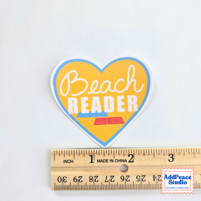 Heart-shaped sticker with the words 'Beach READER' in white script and block lettering over a yellow background, outlined in blue, shown above a wooden ruler for scale on a white background.