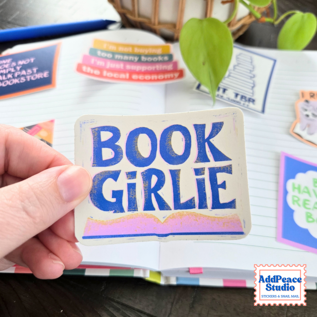 A hand holds a rectangular sticker that reads 'BOOK GIRLIE' in blue lettering with a pink and gold wavy line at the bottom, set against an open notebook filled with colorful sticky notes and books.