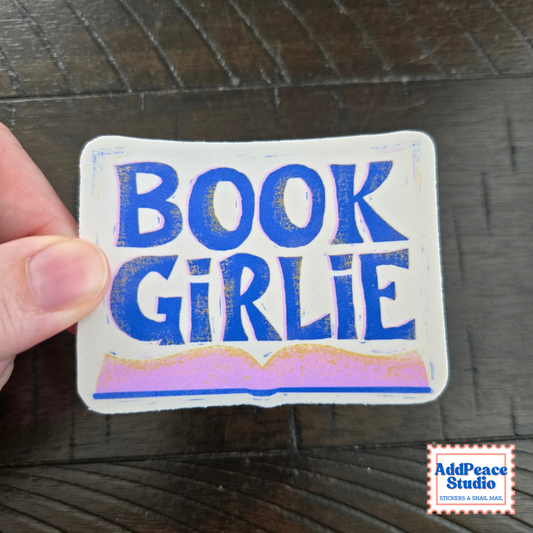 A hand holds a white rectangular sticker with the words 'BOOK GIRLIE' in bold blue letters and a pink and gold open-book illustration at the bottom, set against a dark wood background. An 'AddPeace Studio' logo is visible in the bottom right corner.