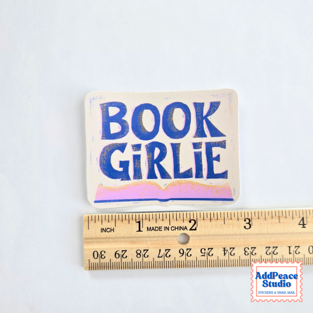 A rectangular fabric sticker with the words 'BOOK GIRLIE' in blue lettering on a cream background, shown above a wooden ruler to indicate scale. The sticker has a small hole at the top for hanging.