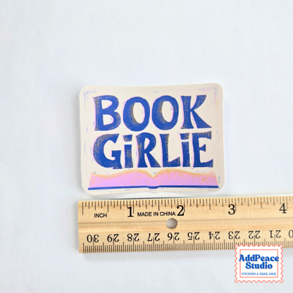 A rectangular fabric sticker with the words 'BOOK GIRLIE' in blue lettering on a cream background, shown above a wooden ruler to indicate scale. The sticker has a small hole at the top for hanging.