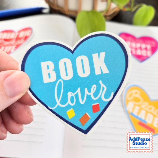 A hand holds a heart-shaped blue sticker that reads 'BOOK lover' in white lettering, with small colorful book icons near the bottom. The background shows an open notebook with other heart-shaped stickers and a blurred plant.