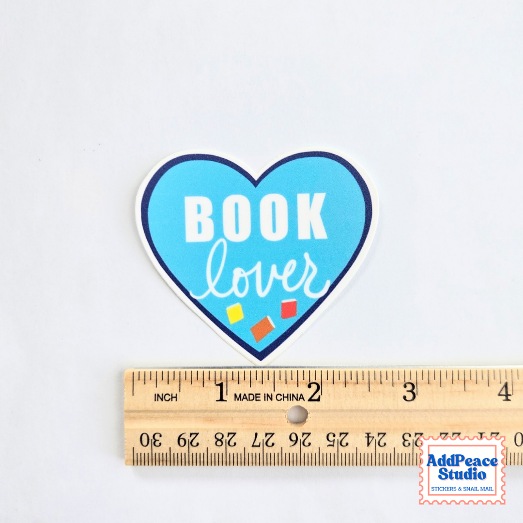 A heart-shaped sticker with the words 'BOOK lover' in white on a light blue background, shown above a wooden ruler for scale on a white surface.