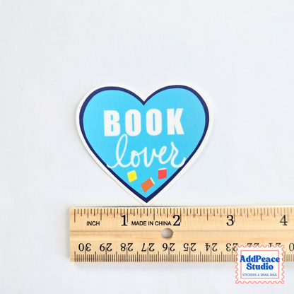 A heart-shaped sticker with the words 'BOOK lover' in white on a light blue background, shown above a wooden ruler for scale on a white surface.