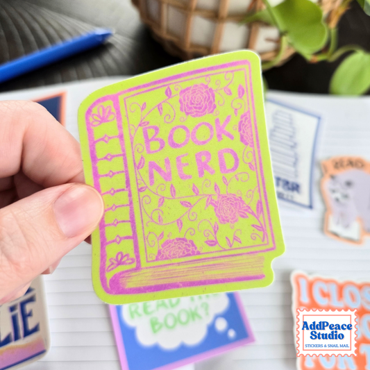 A hand holds a die-cut sticker shaped like a book with the words 'BOOK NERD' in pink on a bright yellow-green background. Other colorful stickers and reading-themed items are blurred in the background, with an 'AddPeace Studio' logo in the bottom right corner.