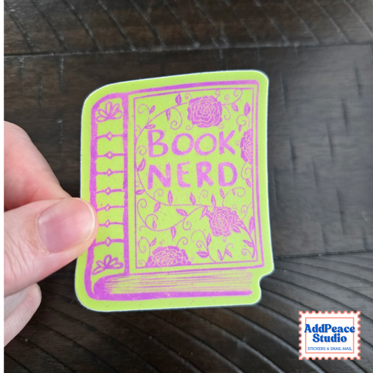 A hand holds a die-cut sticker shaped like a book with the words 'BOOK NERD' in pink on a bright yellow-green background. The sticker is photographed over a dark wood surface, with an AddPeace Studio logo in the bottom right corner.