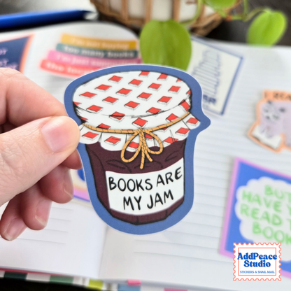 A hand holds a die-cut sticker shaped like a jar of jam with the text 'BOOKS ARE MY JAM'. The sticker has a blue border, a white and red checkered lid, and a brown jam-filled body.