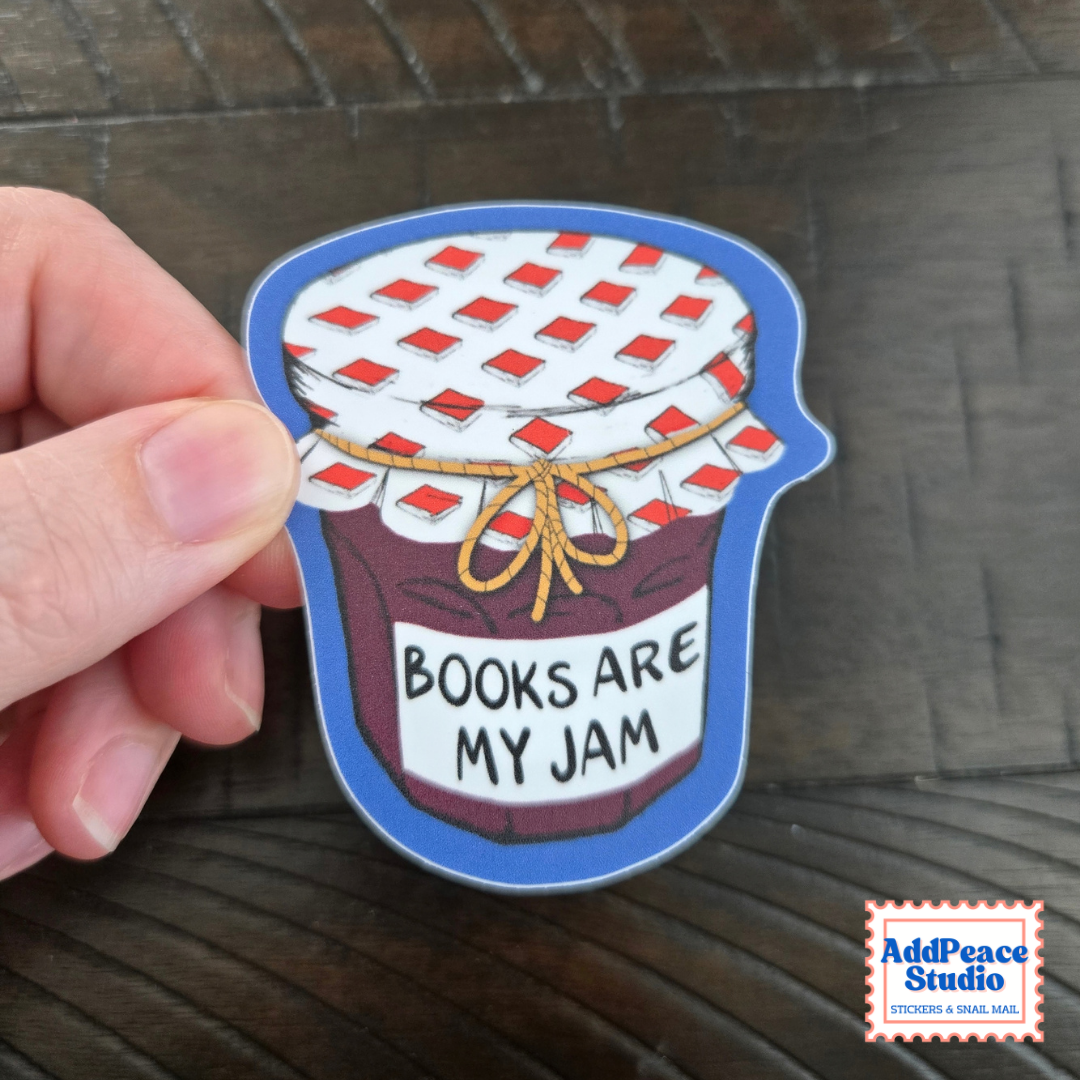 A hand holds a die-cut sticker shaped like a jar of jam with the text 'BOOKS ARE MY JAM' on a dark wooden surface. The sticker has a blue border, a white lid with red patterned squares, brown jam, and a yellow twine bow.