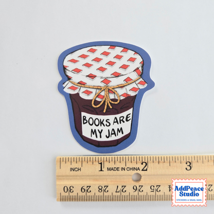 A die-cut sticker shaped like a jar of jam with the text 'BOOKS ARE MY JAM' on a white background, shown above a wooden ruler for scale. The sticker has a blue border, a white lid with red diamond pattern, brown jam, and a beige twine bow.
