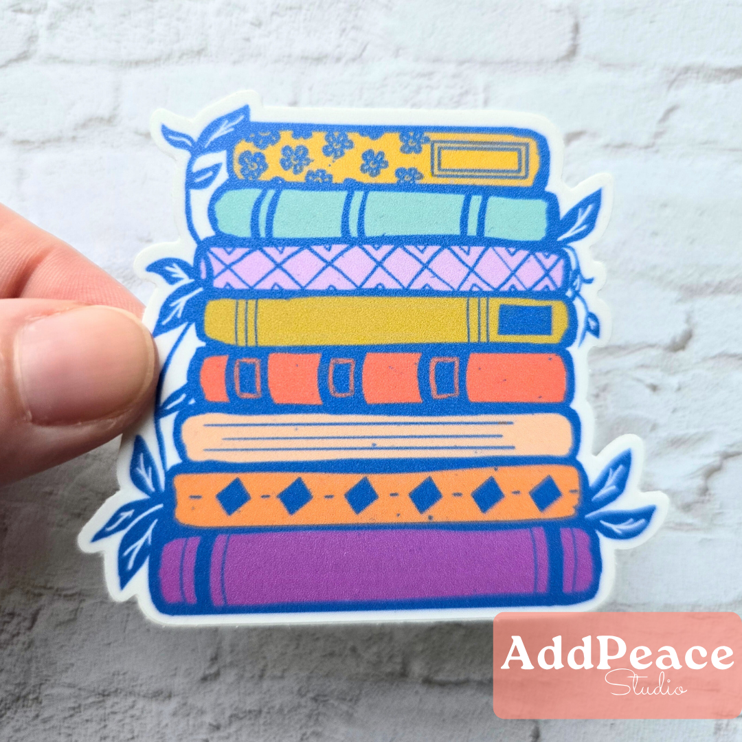 Colorful stack of books sticker held by a hand against a textured white background with 'AddPeace Studios' branding.