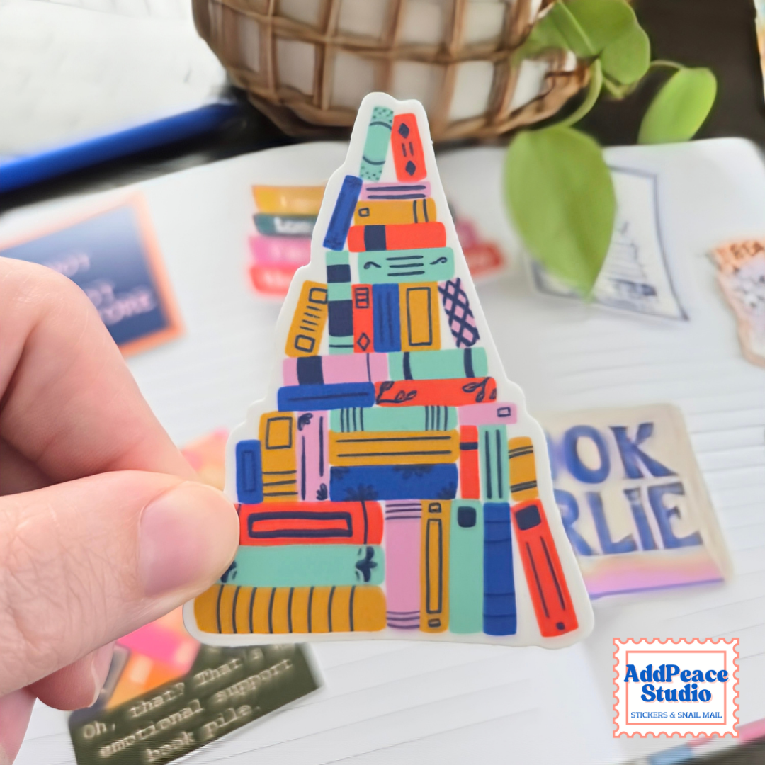 A hand holds a die-cut sticker shaped like a book pile, featuring a colorful pattern of stacked books in teal, orange, pink, yellow, and blue. The background shows a desk with other stickers, a plant, and a basket.
