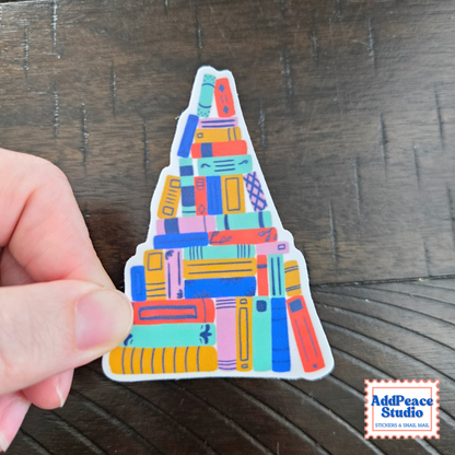 A hand holds a die-cut sticker shaped like a Christmas tree made of colorful stacked books, set against a dark wood background. An 'AddPeace Studio' brand watermark is visible in the bottom right corner.
