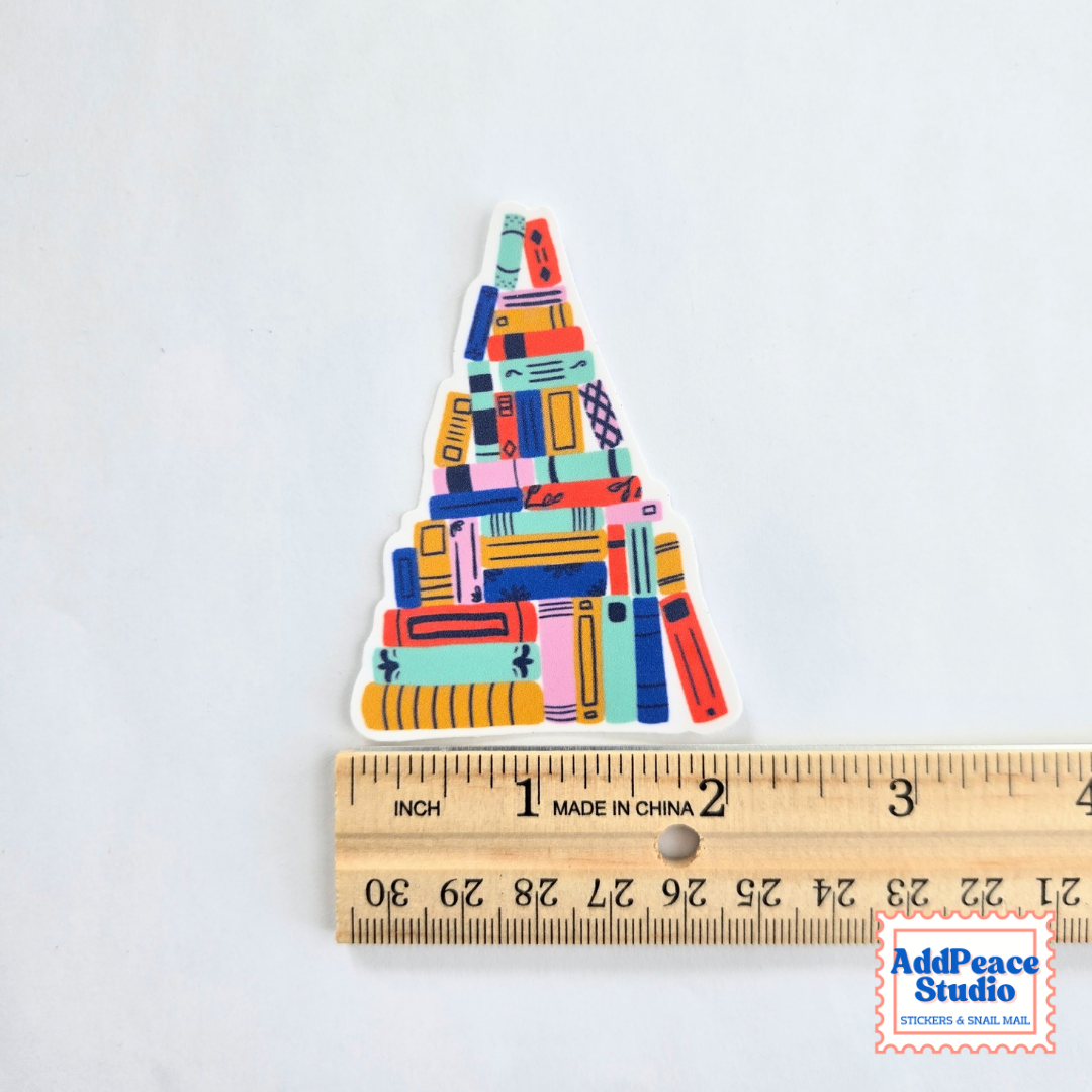 A colorful triangular sticker featuring an illustrated stack of books in various bright colors, shown next to a wooden ruler for scale on a white background.