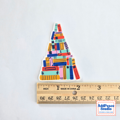 A colorful triangular sticker featuring an illustrated stack of books in various bright colors, shown next to a wooden ruler for scale on a white background.