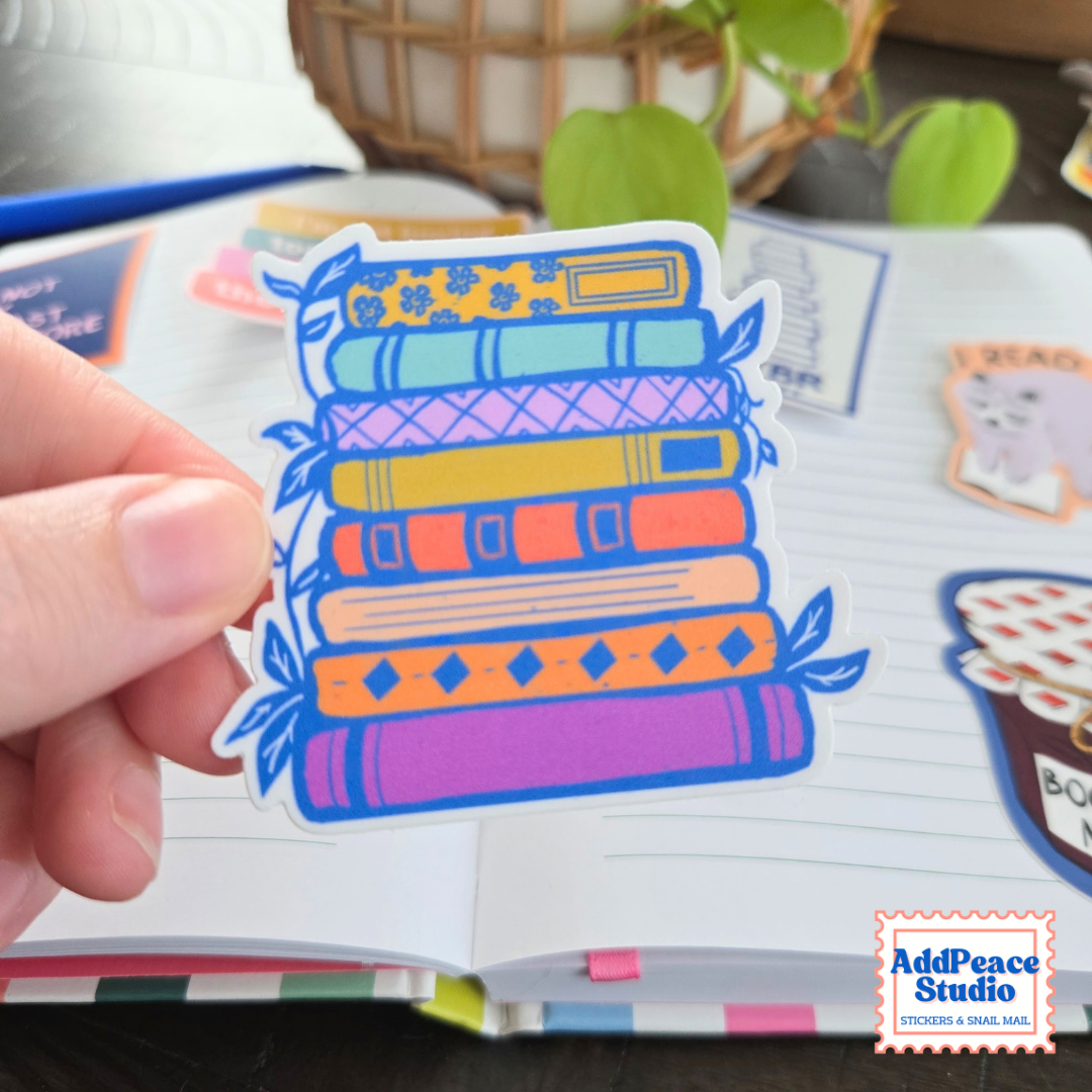 A hand holds a die-cut sticker shaped like a stack of colorful patterned books. The sticker is shown in front of an open notebook with other stickers visible on the page.