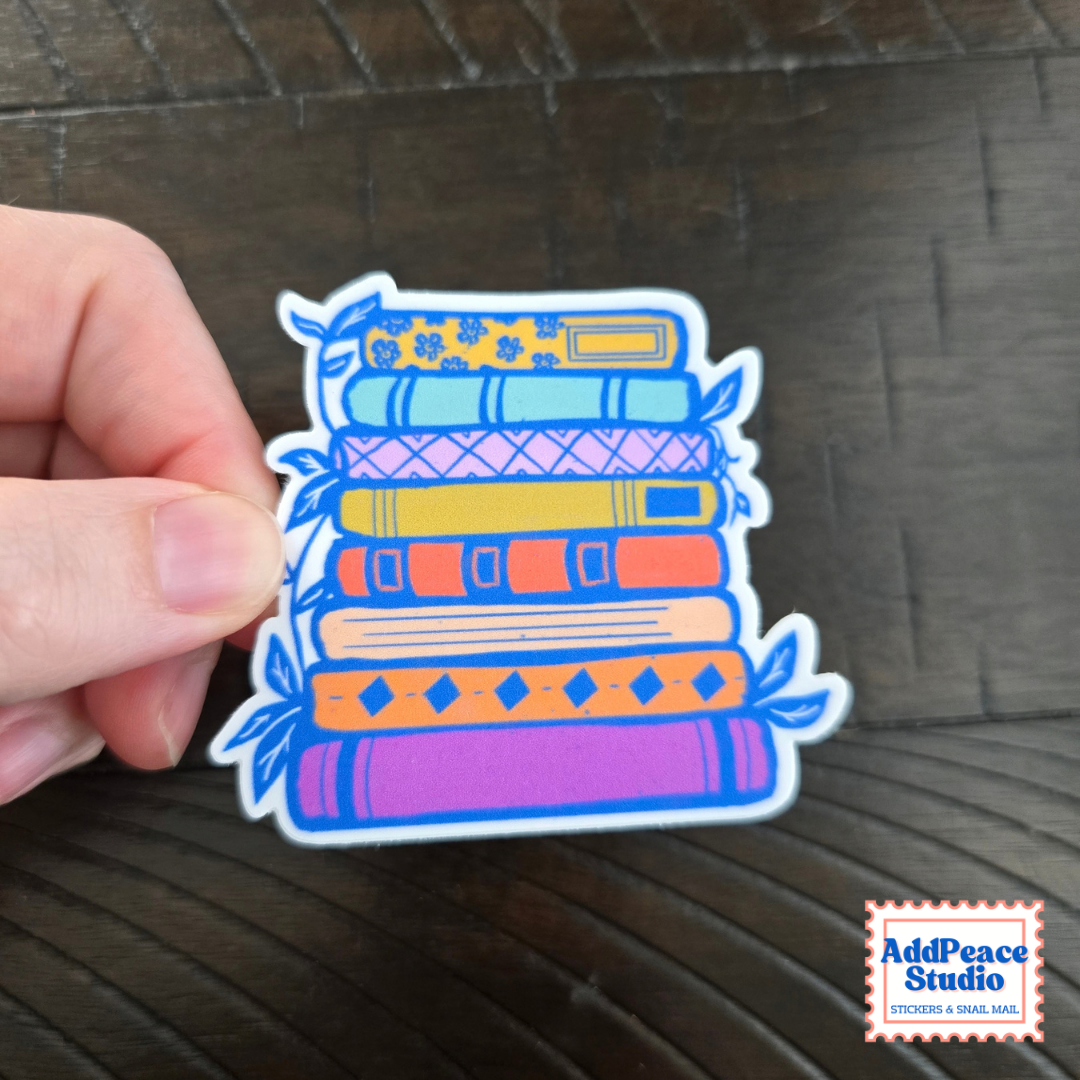 A hand holds a die-cut sticker featuring a colorful stack of books with blue leaves on the sides, set against a dark wood background. The AddPeace Studio logo is visible in the bottom right corner.
