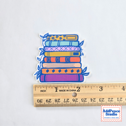 A die-cut sticker depicting a colorful stack of books in shades of blue, purple, orange, and yellow, shown next to a wooden ruler for scale on a white background.