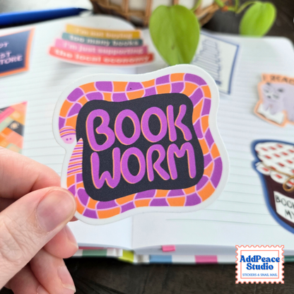 A hand holds a die-cut sticker featuring the words 'BOOK WORM' in pink lettering on a black background, surrounded by a purple and orange illustrated worm. The sticker is shown against a blurred background of lined journal pages and other stickers, with a 'AddPeace Studio' logo in the bottom right corner.