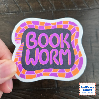 A hand holds a die-cut sticker featuring the words 'BOOK WORM' in pink lettering on a dark gray background, surrounded by a purple and orange illustrated worm. The sticker is photographed on a dark wood surface, with an 'AddPeace Studio' logo in the bottom right corner.