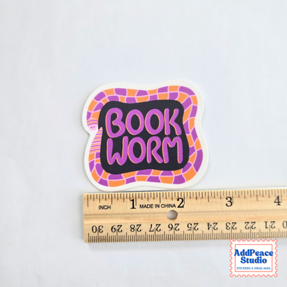 A sticker with purple lettering that says "BOOK WORM" surrounded by a purple and orange illustrated worm on a dark background. A ruler is pictured to show scale.
