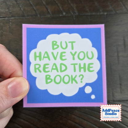 A hand holds a square card with a blue background, a light purple border, and a white thought bubble containing the green text 'BUT HAVE YOU READ THE BOOK?'. A small AddPeace Studio logo appears in the bottom right corner, with a dark wood surface in the background.