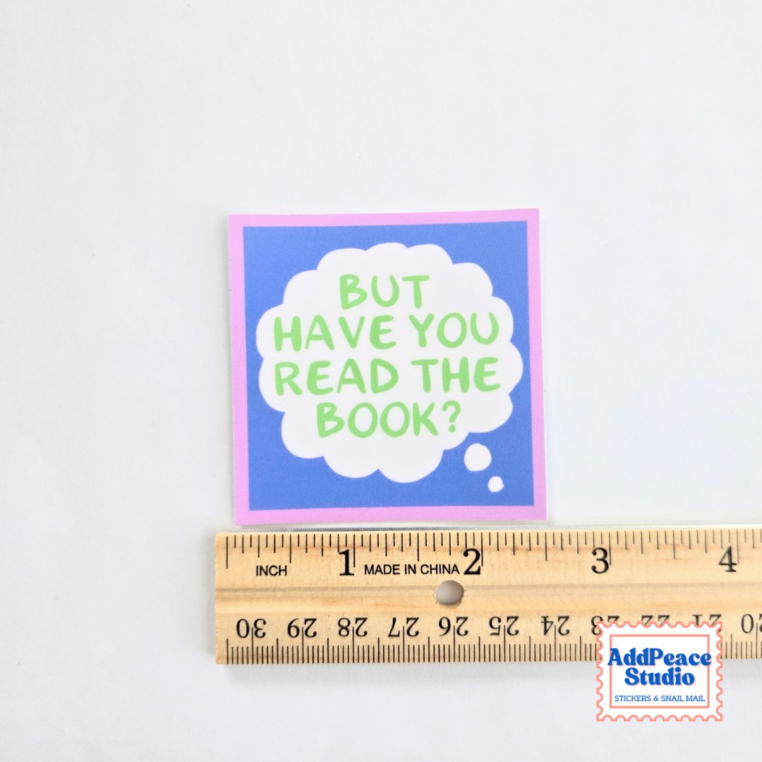 A square sticker with a blue center, pink border, and a white thought-bubble shape containing the phrase “BUT HAVE YOU READ THE BOOK?” in light green letters, shown above a wooden ruler on a white background. An AddPeace Studio logo appears in the bottom right corner.