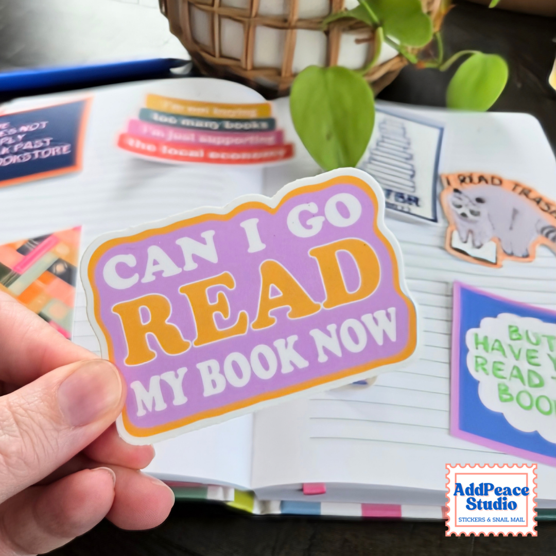 A hand holds a die-cut sticker that reads 'CAN I GO READ MY BOOK NOW' in purple, orange, and white. The sticker is shown in front of an open book with colorful bookmarks, with a plant in the background.