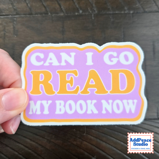 A hand holds a die-cut sticker that reads 'CAN I GO READ MY BOOK NOW' in white and orange lettering on a lavender background, with a white and orange border. The sticker is photographed over a dark wood surface, and an 'AddPeace Studio' logo is visible in the bottom right corner.