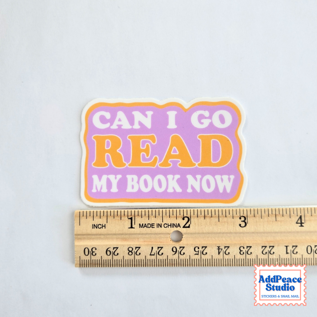 A die-cut sticker with the text 'CAN I GO READ MY BOOK NOW' in white and orange lettering on a lavender and orange background, shown above a wooden ruler for scale on a white background.