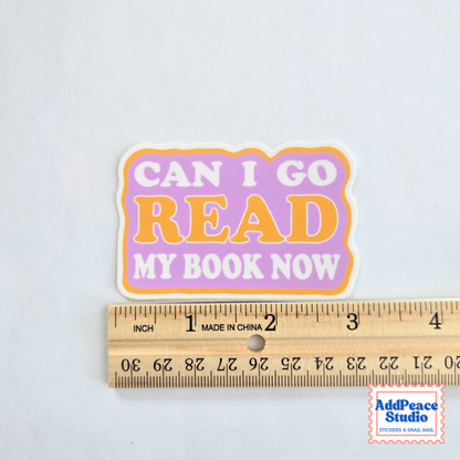 A die-cut sticker with the text 'CAN I GO READ MY BOOK NOW' in white and orange lettering on a lavender and orange background, shown above a wooden ruler for scale on a white background.