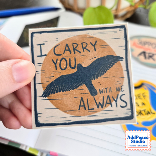 A hand holds a square printed sticker with the words “I CARRY YOU WITH ME ALWAYS” and a dark blue bird silhouette over a tan circle on a cream background. The scene is a real desk environment with other stickers and objects blurred in the background, so this is Lifestyle rather than Product/Packaging.