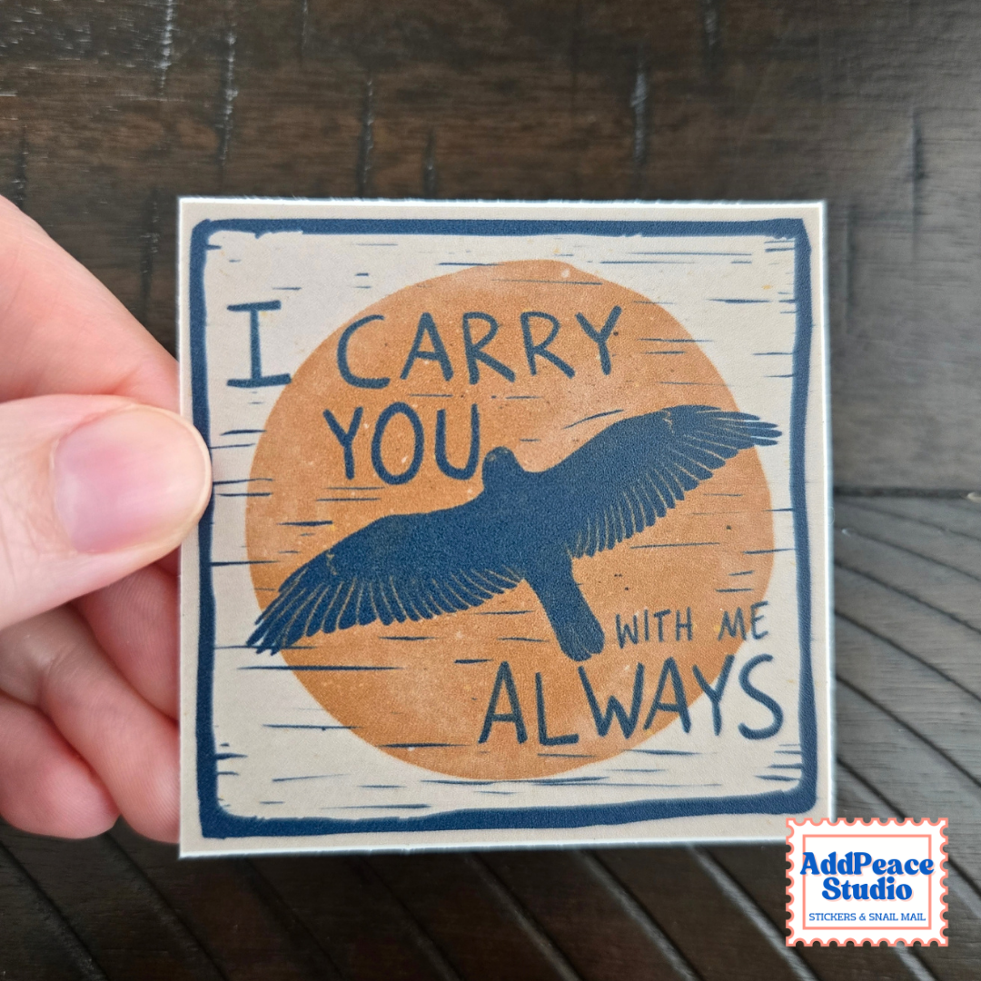 A hand holds a square sticker with a navy blue bird over an orange circle and the words “I CARRY YOU WITH ME ALWAYS” on a dark wood surface. A small AddPeace Studio logo appears in the bottom right corner.