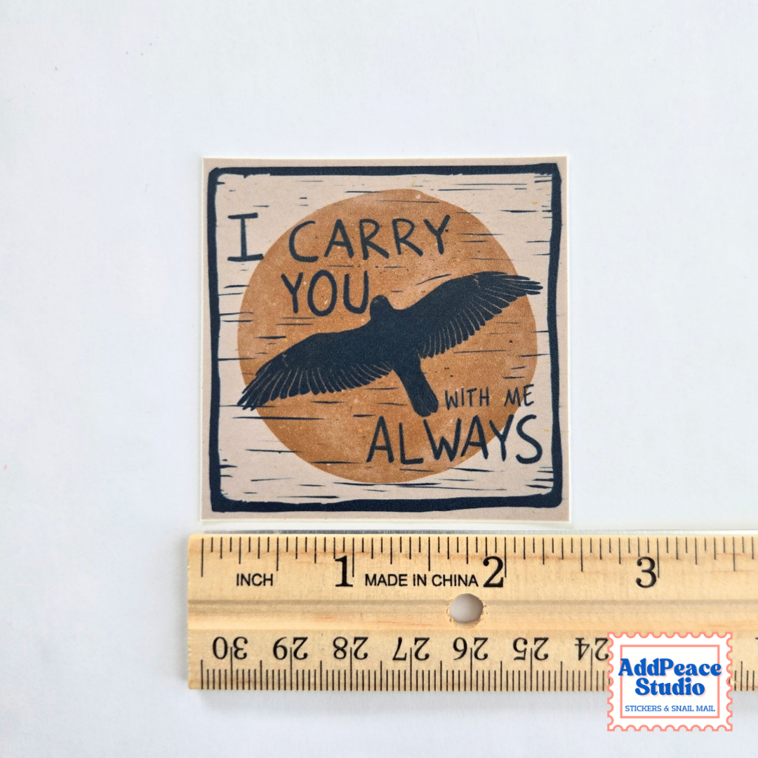 A square printed sticker or decal with the phrase “I CARRY YOU WITH ME ALWAYS” and a black bird silhouette over a tan circle, shown above a wooden ruler for scale on a white background.