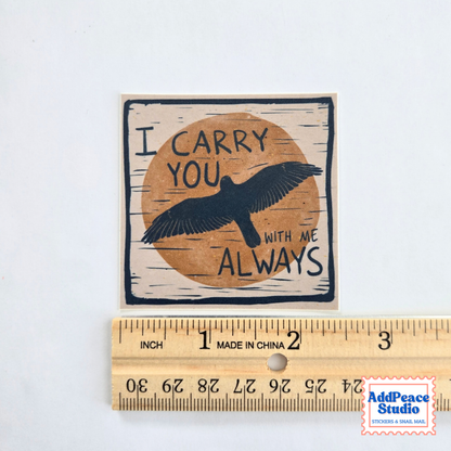 A square printed sticker or decal with the phrase “I CARRY YOU WITH ME ALWAYS” and a black bird silhouette over a tan circle, shown above a wooden ruler for scale on a white background.