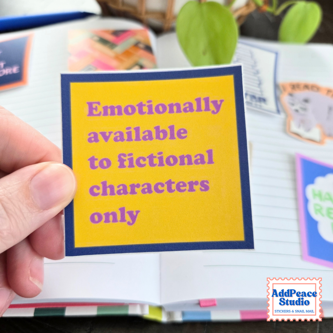 A hand holds a square yellow sticker with a navy blue border and purple text that reads 'Emotionally available to fictional characters only.' The background shows an open notebook with other colorful stickers and a blurred plant.