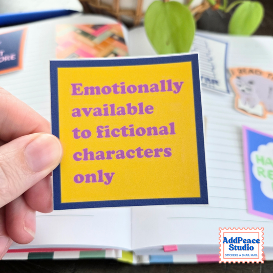 A hand holds a square yellow sticker with a navy blue border and purple text that reads 'Emotionally available to fictional characters only.' The background shows an open notebook with other colorful stickers and a blurred plant.