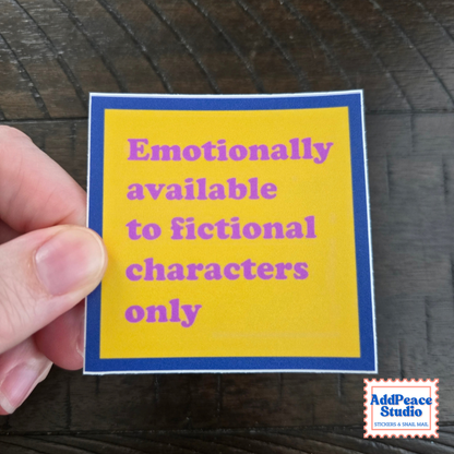 A hand holds a square yellow sticker with a blue border and purple text that reads 'Emotionally available to fictional characters only' against a dark wood background. AddPeace Studio logo is visible in the bottom right corner.