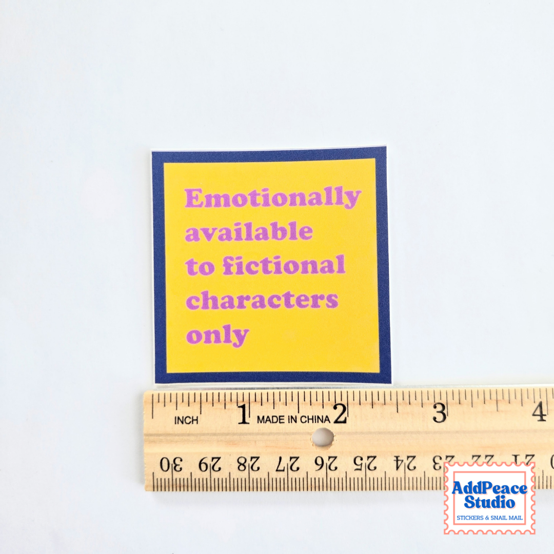 A square yellow sticker with a dark blue border and pink text reading 'Emotionally available to fictional characters only,' shown above a wooden ruler for scale on a white background.