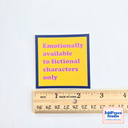 A square yellow sticker with a dark blue border and pink text reading 'Emotionally available to fictional characters only,' shown above a wooden ruler for scale on a white background.