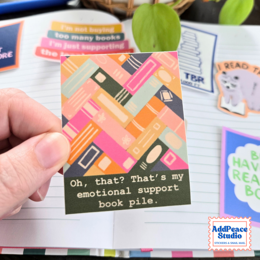A hand holds a small sticker with a colorful illustrated pile of books and the text, "Oh, that? That's my emotional support book pile." The background shows lined paper and other decorative stickers or cards, with a visible AddPeace Studio logo in the bottom right corner.